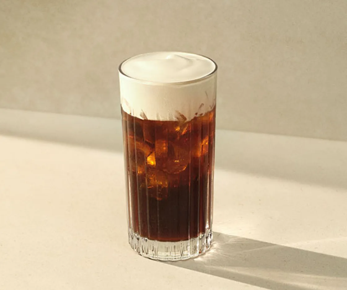 Imagen de un vaso de cristal, dentro tiene un cafe iced americano con espuma de proteína