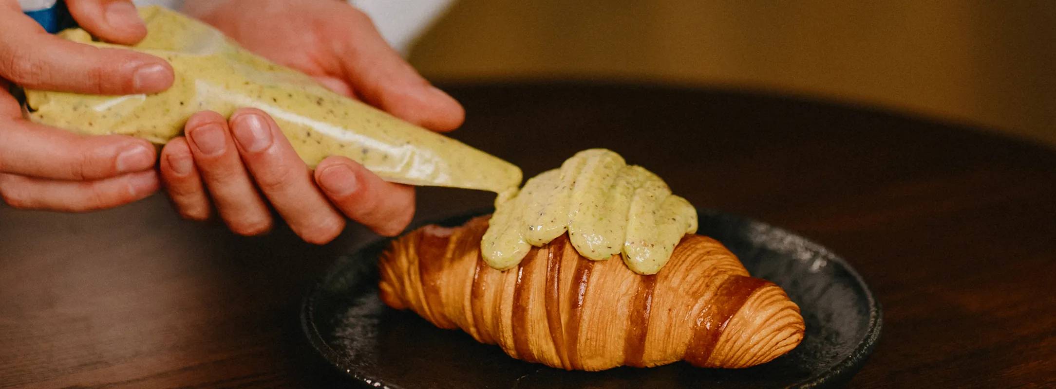 En la imagen se ve un croissant siendo terminado por las manos de un cocinero, sobre plato negro