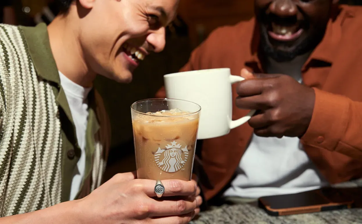 Dos clientes de Starbucks disfrutan de sus bebidas mientras se ríen. Uno tiene un su mano una bebida con hielo en vaso de cristal y otro una bebida caliente en la icónica taza blanca de Starbucks